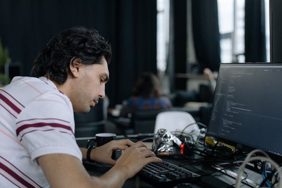 A young man with dark, wavy hair, wearing a white t-shirt with red and purple stripes, is seated at a desk in an office or classroom environment, focused on typing on a black keyboard. In front of him
